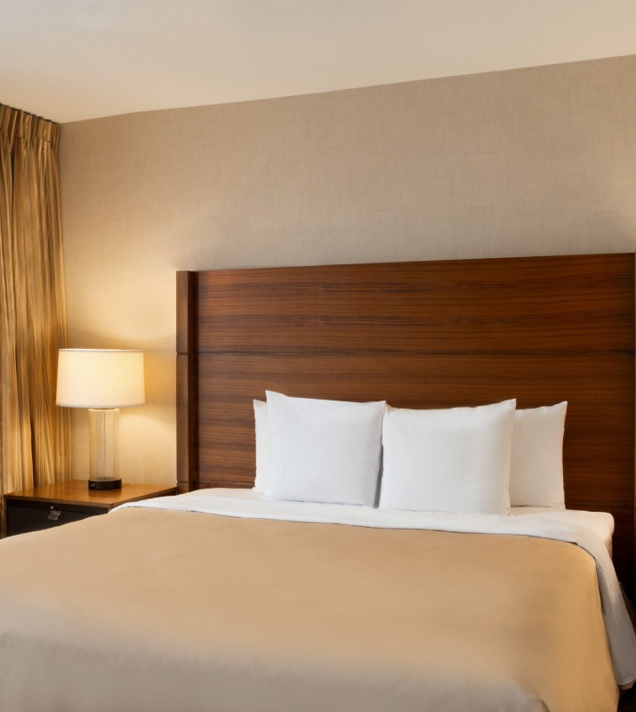 King size hotel room with warm lighting, dark wood, and beige bedding inside of Homewood Suites in Harbor East