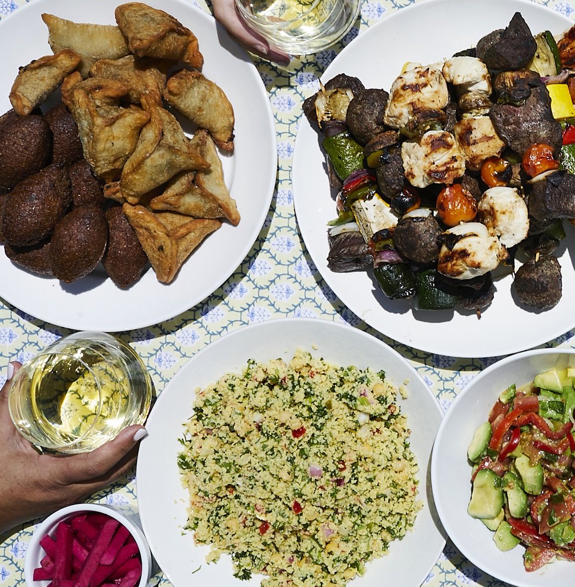 Overhead table spread of various mediterranean-themed foods and sides, with hands reaching in holding glasses of white wine