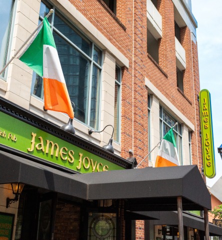 Closeup exterior of James Joyce Pub featuring brick storefront, black covered awning, and Irish flags