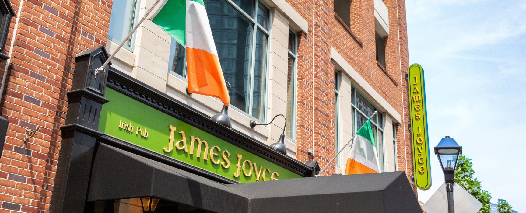 Wide angle closeup exterior of James Joyce Pub featuring brick storefront, black covered awning, and Irish flags