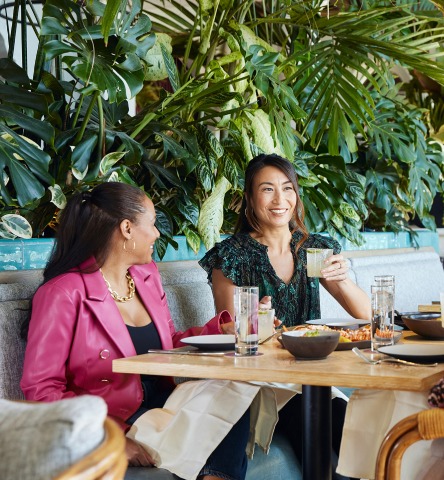 Two women dining over cocktails at a booth inside Maximon restaurant surrounded by greenery