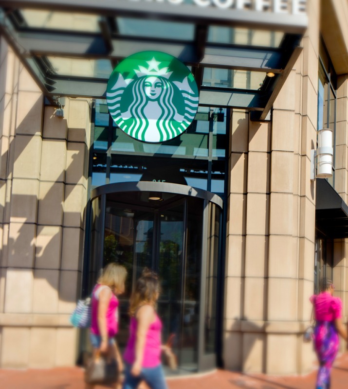 Exterior of Starbucks cafe with green logo in focus and shoppers walking by that are blurred