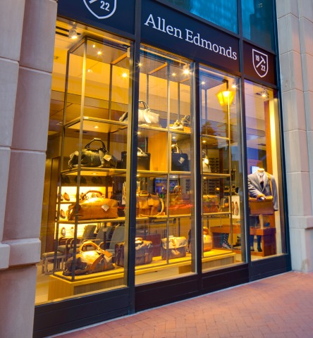 Allen Edmonds storefront, featuring several travel bags and upscale suits at twilight