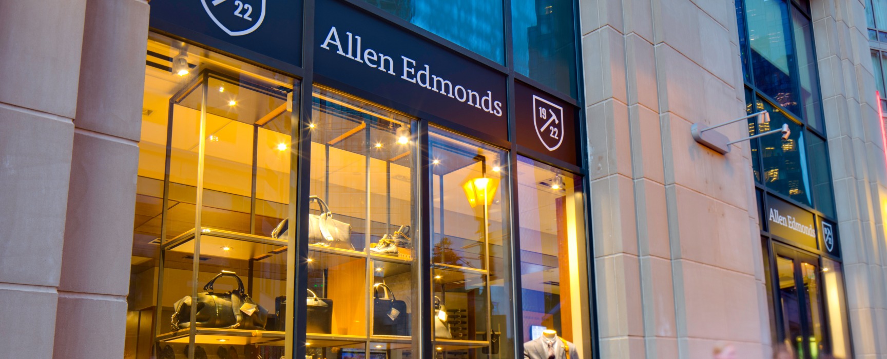 Allen Edmonds storefront, featuring several travel bags and upscale suits at twilight