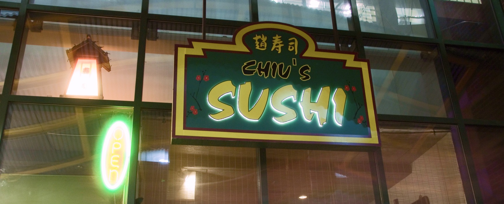 Green and yellow exterior business sign of Chiu's Sushi, lit up and on the exterior window of the storefront