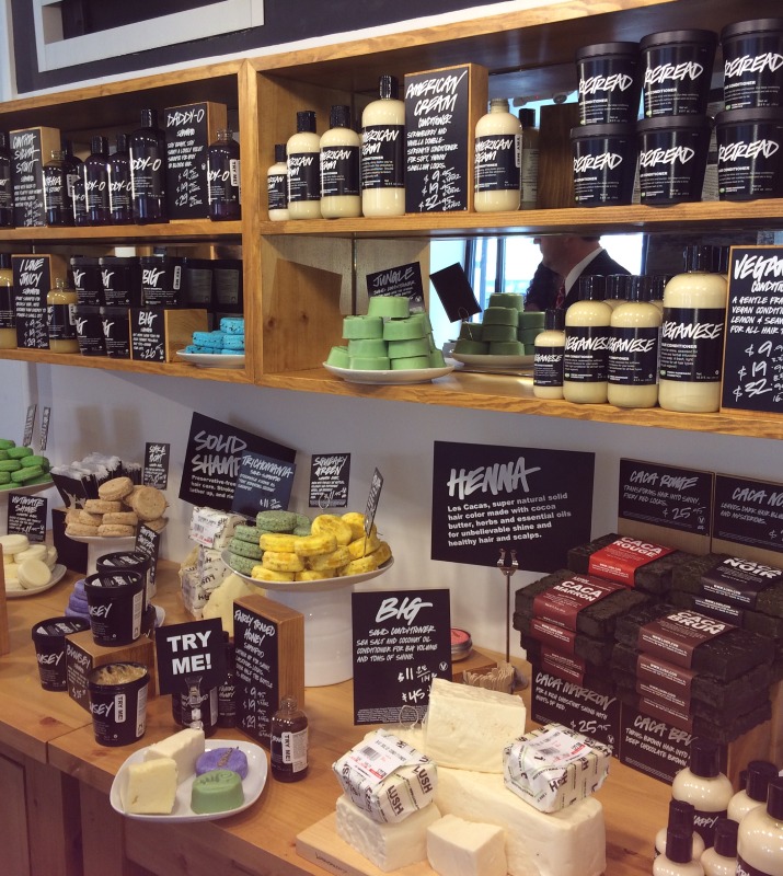 Several soaps, hair products, creams, and other cosmetics are displayed on light wood shelves in Lush
