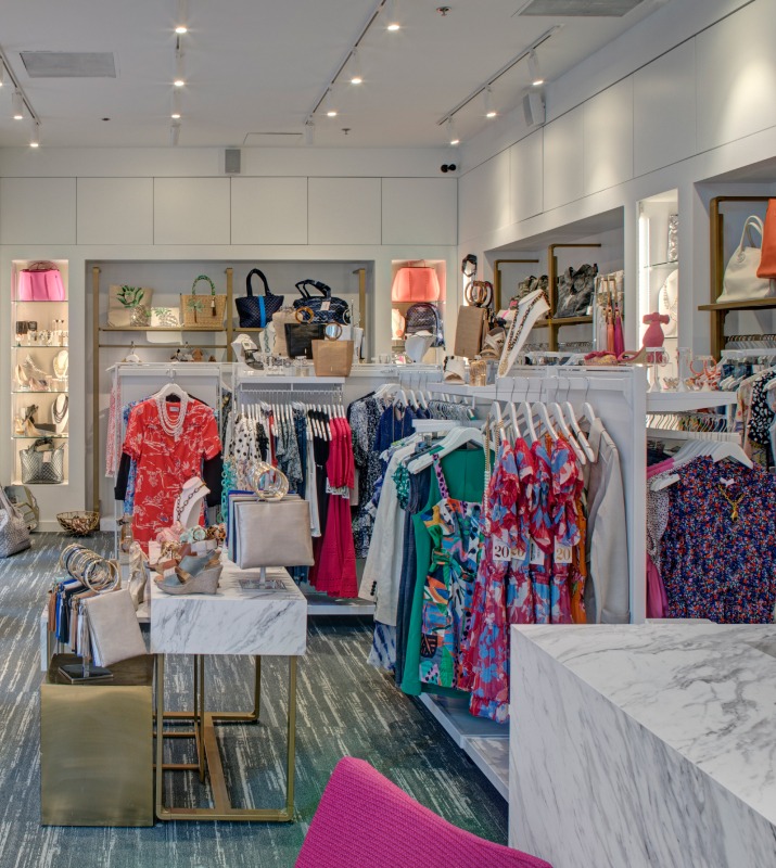 Brightly colored dresses, shirts, and other clothing displayed on racks with accessories on shelves inside of Sassanova boutique in Harbor East