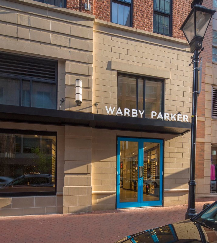 Exterior brick and block storefront of Warby Parker with blue frame window door and black awning to hold the store sign