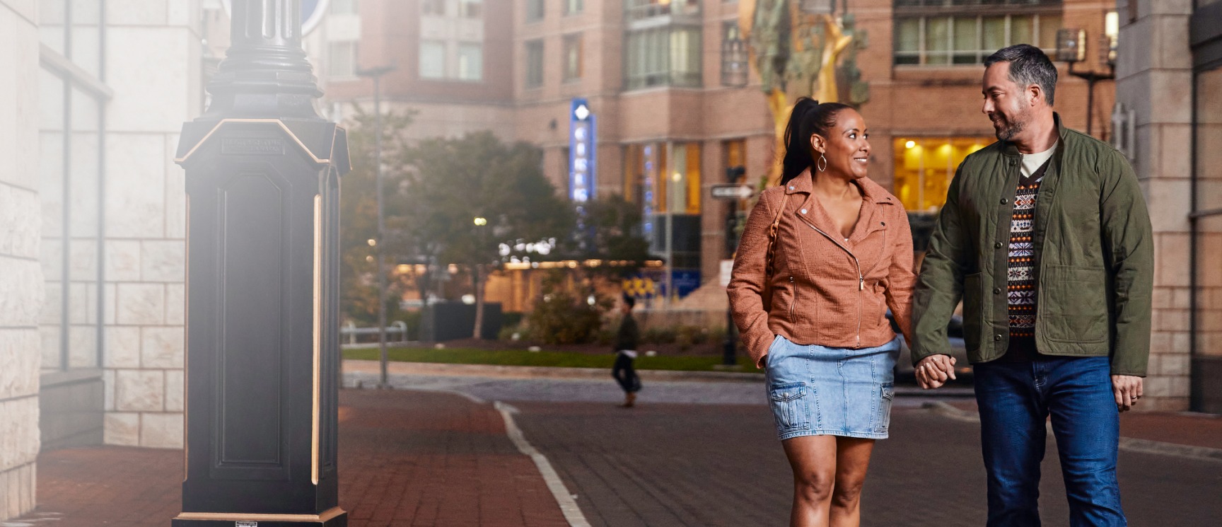 A man and a woman hold hands as they walk through the center of Harbor East at dusk