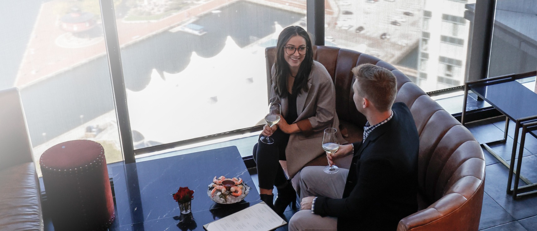 A man and a woman share drinks and shrimp cocktail inside of a booth at The Bygone with a view of Baltimore City from above