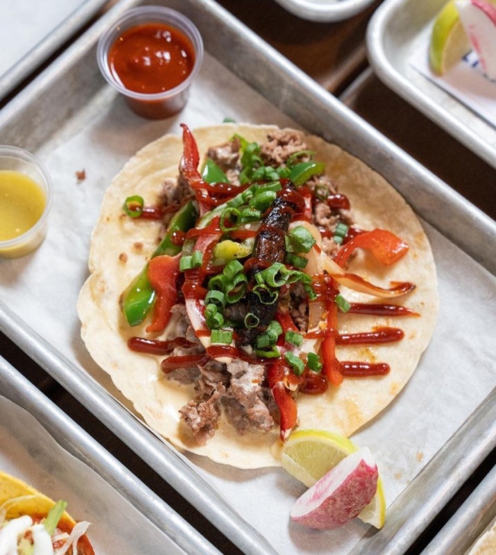 Overhead view of open taco on metal tray featuring meat, various colorful toppings, and neatly drizzled sauces