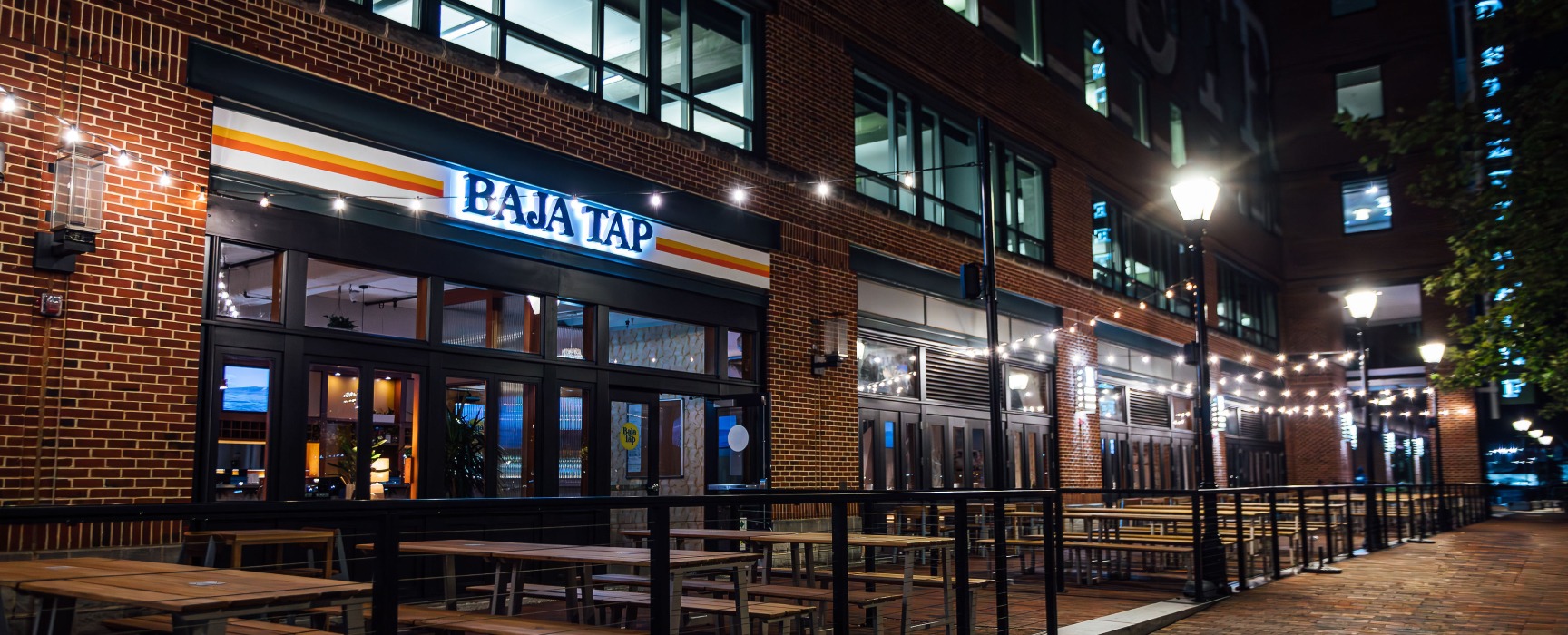 Exterior of Baja Tap restaurant brick storefront at night with illuminated sign and outdoor tables